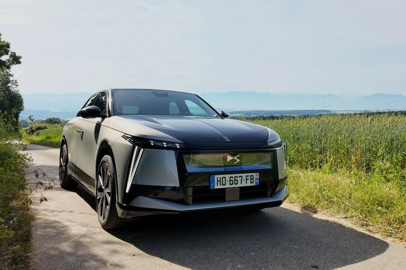 Luksusowy samochód osobowy DS w nowoczesnej stylistyce, zaparkowany na wąskiej drodze w otoczeniu zielonych pól, z panoramicznym widokiem na odległe wzgórza.