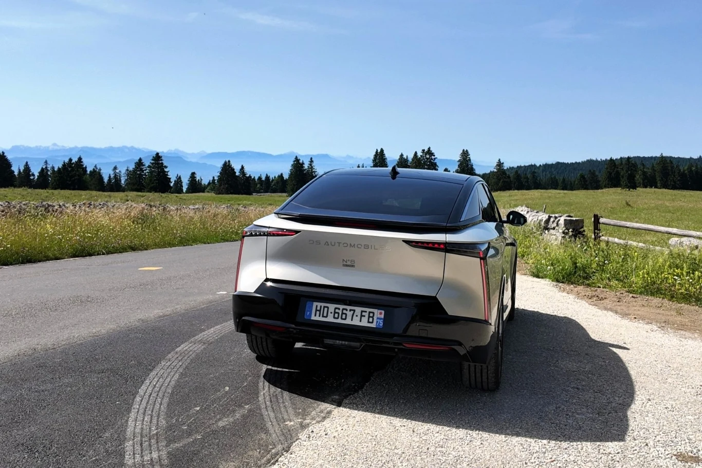Srebrny samochód elektryczny DS Automobiles zaparkowany przy bocznej drodze wśród zielonych łąk, w tle górzysty krajobraz i linia lasu, wyraźna tablica rejestracyjna pojazdu, ślady opon na asfalcie.