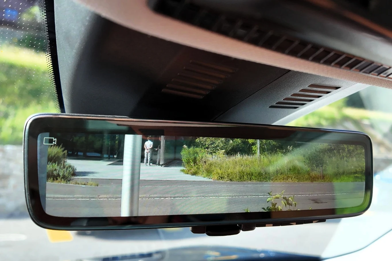 Lusterko wsteczne samochodu odbijające widok na zewnętrzną ulicę z zielenią oraz znakiem drogowym na poboczu, wewnątrz pojazdu widoczne także elementy kokpitu.