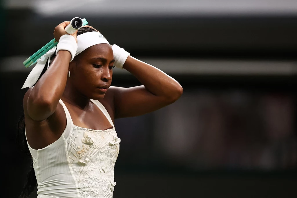 Coco Gauff rywalizowała z Dajaną Jastremską w pierwszej rundzie Wimbledonu Tenisistka w białym stroju sportowym poprawia opaskę na głowie, trzymając rakietę tenisową, wyrażając koncentrację i determinację.