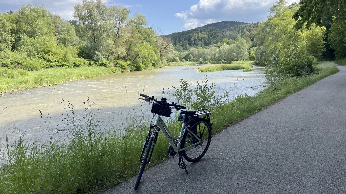 Rower stojący na asfaltowej ścieżce rowerowej przy malowniczej rzece, otoczonej bujną zielenią i lasem. W tle lekkie wzgórza oraz niebieskie niebo z delikatnymi chmurami.
