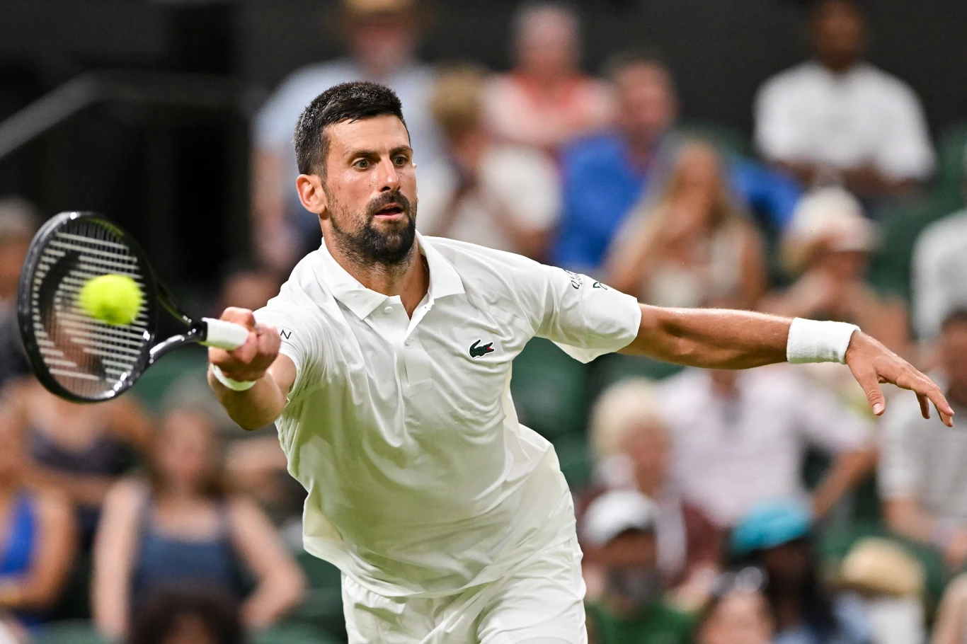 Tenisista w białym stroju podczas dynamicznego ruchu zamachowego do piłki na korcie, wokół widać rozmytą publiczność na trybunach.