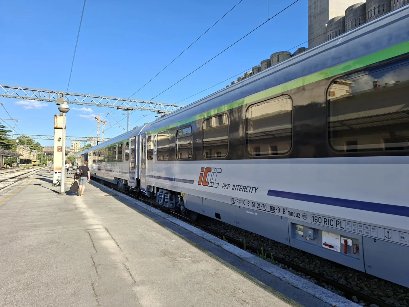 Peron kolejowy z nowoczesnym pociągiem PKP Intercity, jasne wagony z niebieskim i zielonym paskiem, widoczna infrastruktura stacji oraz kilka osób zmierzających do wagonu