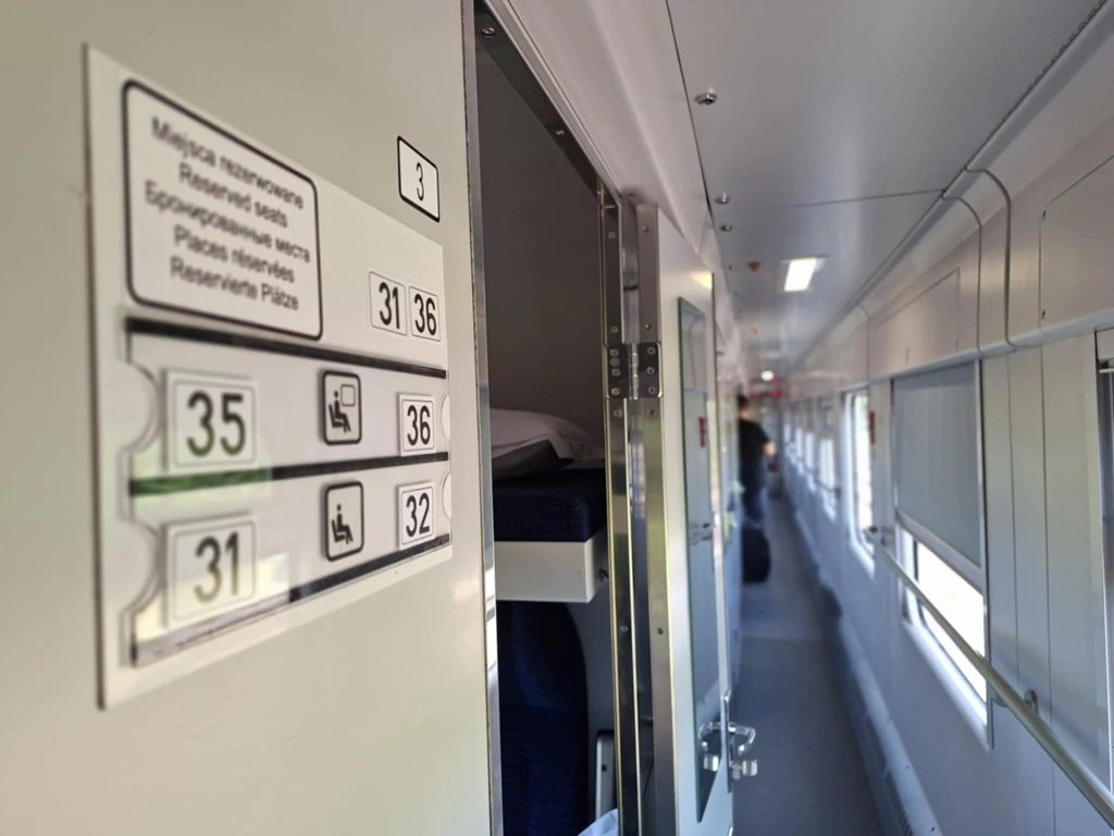 Tabliczka z numerami miejsc siedzących przy wejściu do przedziału w nowoczesnym wagonie kolejowym, światło dzienne wpada przez okna boczne, korytarz wydaje się pusty, panuje spokojna atmosfera.