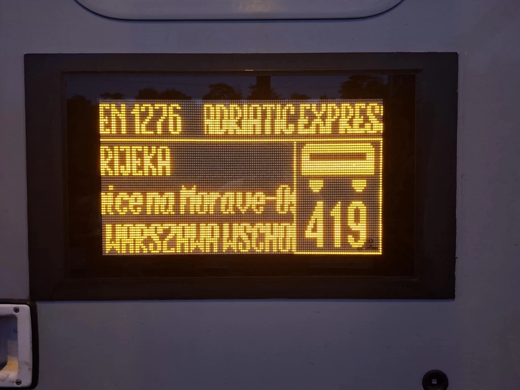Elektroniczny wyświetlacz informacyjny z żółtym napisem, wskazujący numer pociągu EN 1276, trasę ADRIATIC EXPRESS do Rijeki, przesiadkę na autobus oraz numer linii 419.