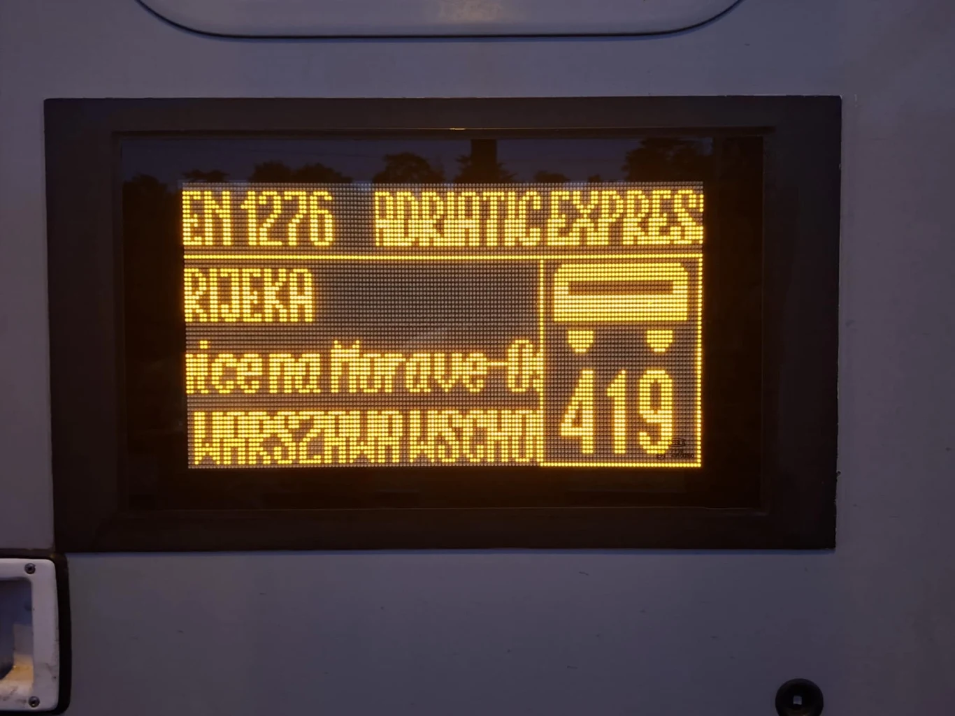 Elektroniczny wyświetlacz informacyjny z żółtym napisem, wskazujący numer pociągu EN 1276, trasę ADRIATIC EXPRESS do Rijeki, przesiadkę na autobus oraz numer linii 419.