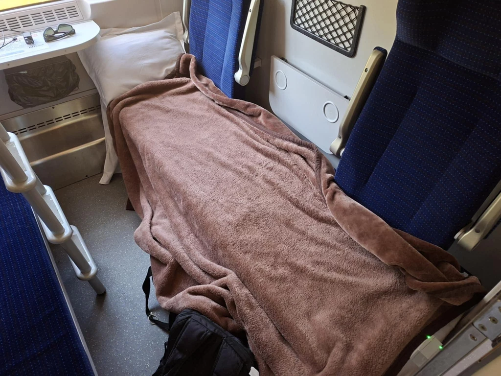 Brązowy koc przykrywa kilka siedzisk w przedziale pociągu, obok znajduje się poduszka oraz niewielki stolik, wnętrze utrzymane jest w niebiesko-szarej tonacji.