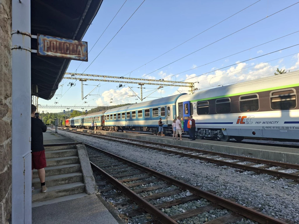 Pociąg pasażerski marki PKP Intercity stoi na peronie stacji kolejowej, kilka osób spaceruje wzdłuż torów, widoczna tablica informacyjna z napisem 'Policja' na budynku po lewej stronie, niebo z chmurami w tle.