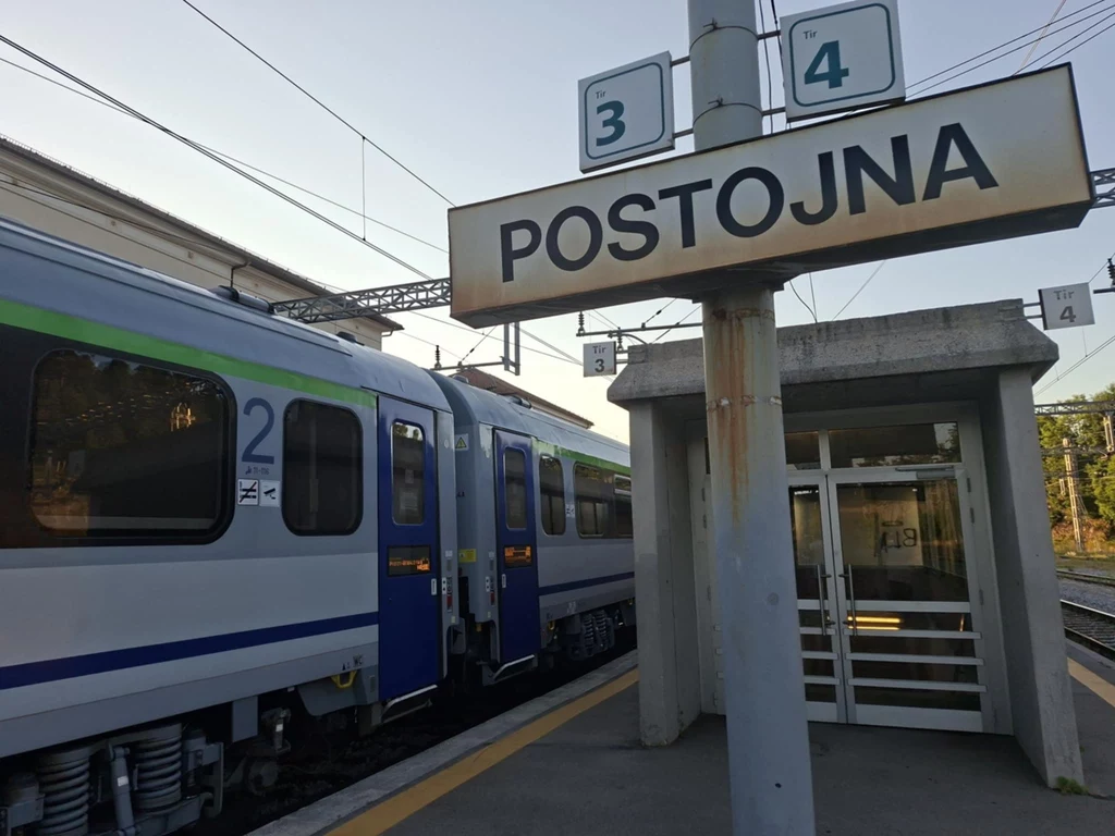 Peron kolejowy z tablicą informacyjną 'POSTOJNA', na którym stoi nowoczesny pociąg pasażerski. Obok peronu znajduje się zadaszone wejście na peron oraz oznaczenia numerów peronów i torów. W tle widoczne są fragmenty infrastruktury kolejowej oraz kabli ...