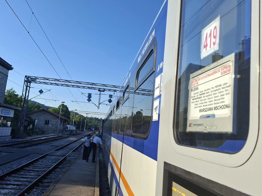 Pociąg pasażerski stoi na bocznicy przy peronie, widoczny rozkład jazdy i numer wagonu, w tle dwie osoby rozmawiają na peronie, niebo bezchmurne, infrastruktura kolejowa z przewodami i słupami.