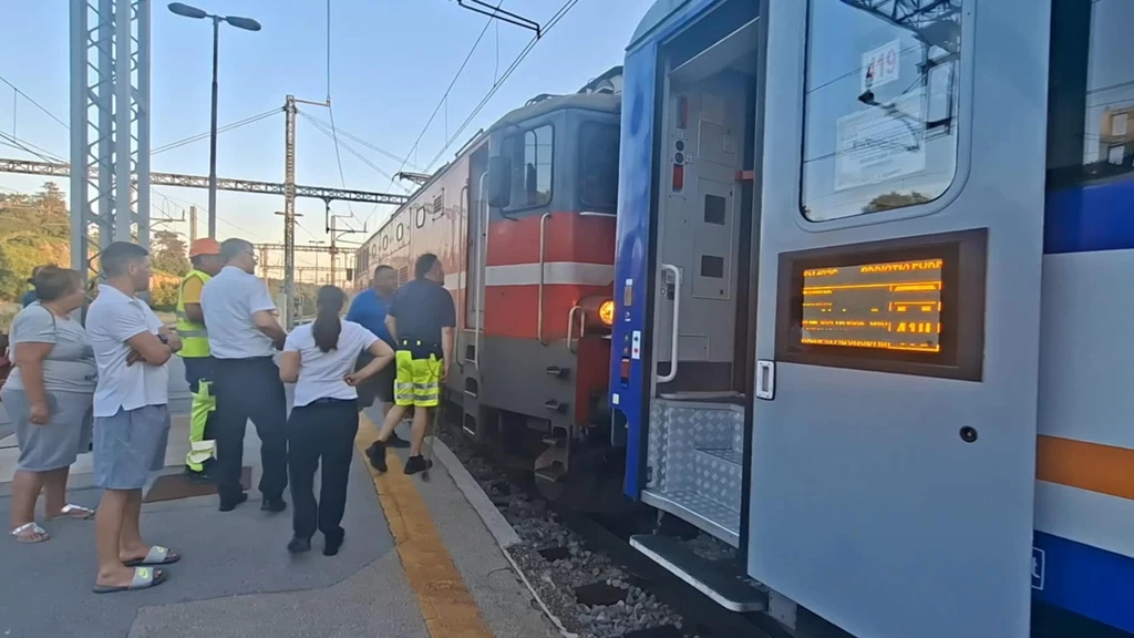 Grupa osób, w tym personel kolejowy i pasażerowie, zgromadzona na peronie obok dwóch pociągów stojących bardzo blisko siebie, przy jednym z pociągów widoczna elektroniczna tablica informacyjna