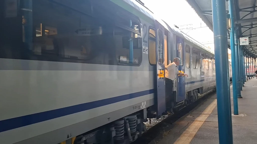 Człowiek w białej koszuli wychylony przez otwarte drzwi wagonu stojącego na peronie kolejowym, wokół spokojna atmosfera stacji, zadaszenie i nieliczne osoby w oddali.