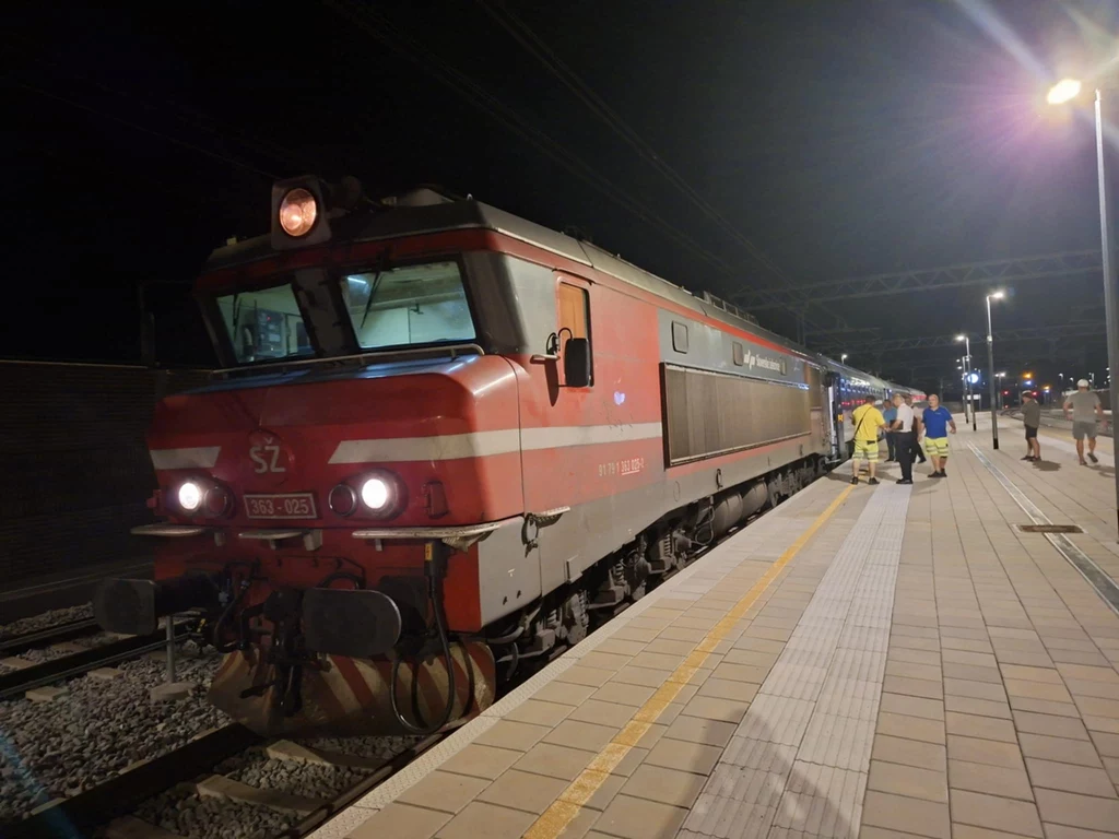Pociąg pasażerski stojący na oświetlonym peronie w nocy, z przodu widoczna lokomotywa z wyraźnym oznaczeniem SZ, kilka osób w jasnych ubraniach znajduje się przy wejściu do wagonów, na dalszym planie rozciąga się peron i latarnie