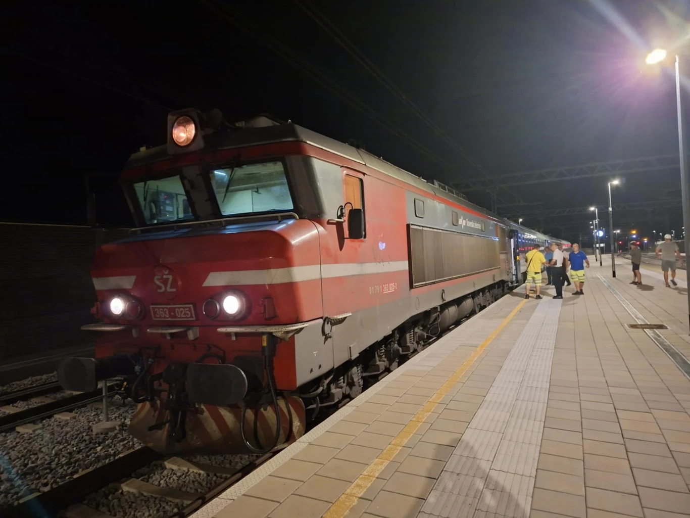 Pociąg pasażerski stojący na oświetlonym peronie w nocy, z przodu widoczna lokomotywa z wyraźnym oznaczeniem SZ, kilka osób w jasnych ubraniach znajduje się przy wejściu do wagonów, na dalszym planie rozciąga się peron i latarnie