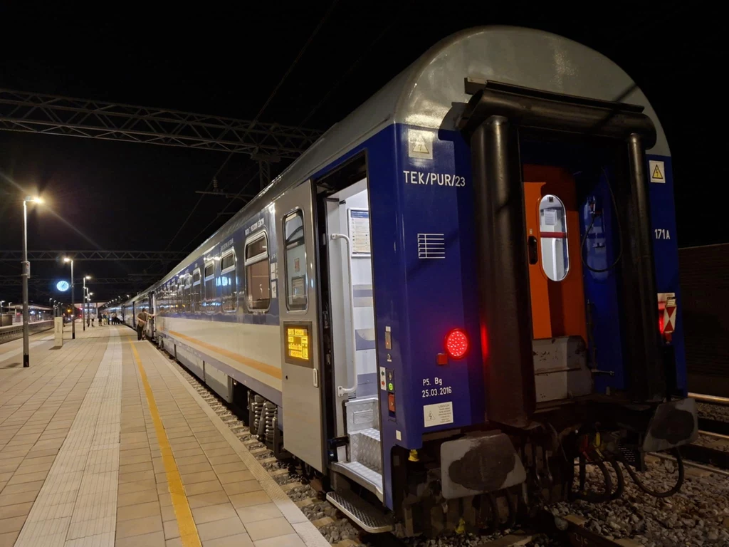 Wagon pociągu pasażerskiego stojący na oświetlonym peronie nocą, widoczne drzwi wejściowe, część infrastruktury kolejowej i napisy informacyjne na wagonie.