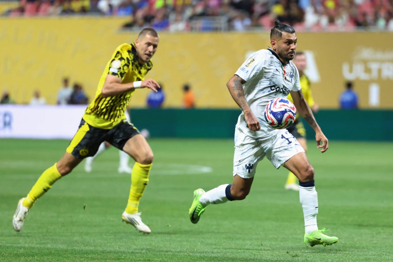 Mecz Borussia Dortmund - Monterrey Dwóch piłkarzy podczas meczu piłki nożnej, zawodnik w białym stroju kontroluje piłkę, a zawodnik w żółto-czarnej koszulce stara się go dogonić, w tle widoczni są kibice na trybunach.