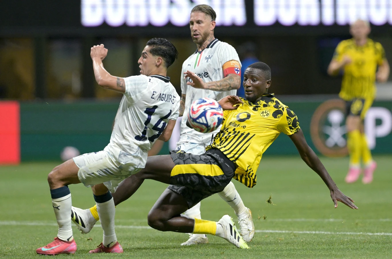 Dwóch piłkarzy walczy o piłkę podczas meczu piłki nożnej, jeden z zawodników w żółto-czarnym stroju wykonuje wślizg próbując odebrać piłkę rywalowi w białym stroju, a w tle widoczny jest kolejny zawodnik.