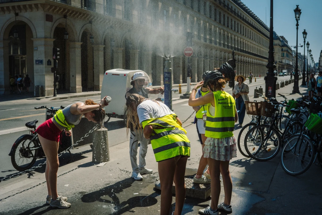 Fala upałów w Europie Grupa osób w kamizelkach odblaskowych korzysta z ulicznego natrysku wodnego w upalny dzień, rowery stoją zaparkowane przy krawężniku, w tle widoczna zabytkowa architektura miasta.