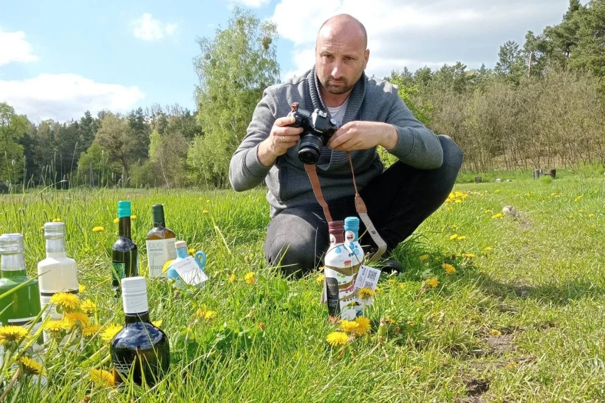Ekspert obala mity związane z oliwą z oliwek Mężczyzna w szarym ubraniu klęczy na trawie i fotografuje rozstawione na trawie butelki z alkoholem oraz inne napoje w otoczeniu kwitnących mleczy, w tle widać las.