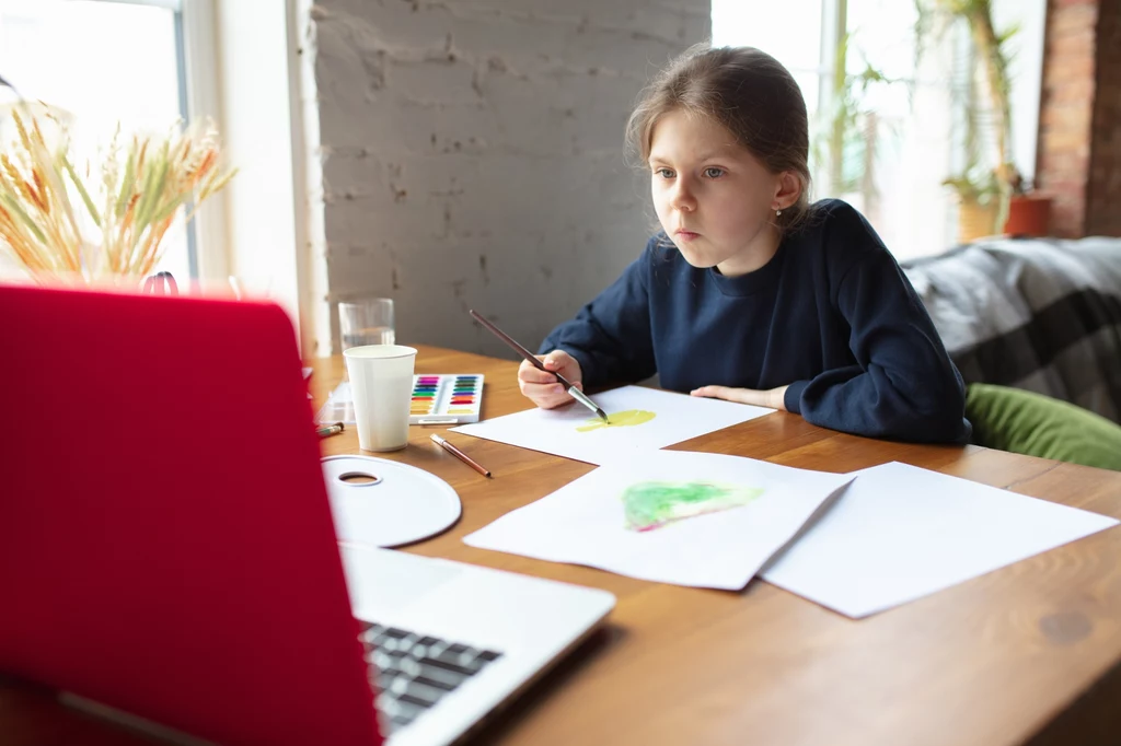 Dziewczynka siedzi przy drewnianym stole, maluje farbami na kartkach papieru, obok niej stoi otwarty laptop, na stole znajdują się także kubek z wodą i przybory plastyczne