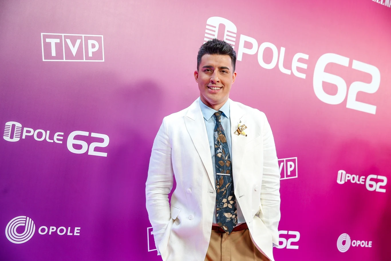 Filip Lato Mężczyzna w jasnej marynarce, niebieskiej koszuli i ozdobnym krawacie stoi na tle ścianki z logotypami TVP oraz Opole 62, uśmiecha się do obiektywu.