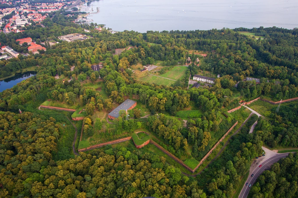 Historyczny fort otoczony gęstym lasem, wyraźnie widoczne ceglane umocnienia w kształcie gwiazdy, liczne budynki oraz rozbudowane ścieżki wśród roślinności, w tle widoczne zabudowania miejskie oraz brzeg jeziora.