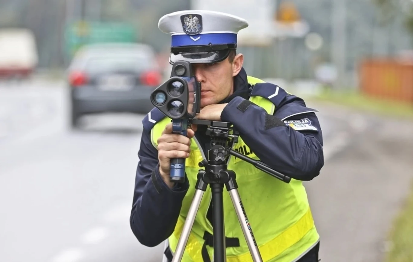 Policjant drogowy w żółtej kamizelce i białej czapce mierzy prędkość przejeżdżających samochodów za pomocą ręcznego radaru ustawionego na statywie, tło stanowi rozmyta droga i pojazdy w ruchu.