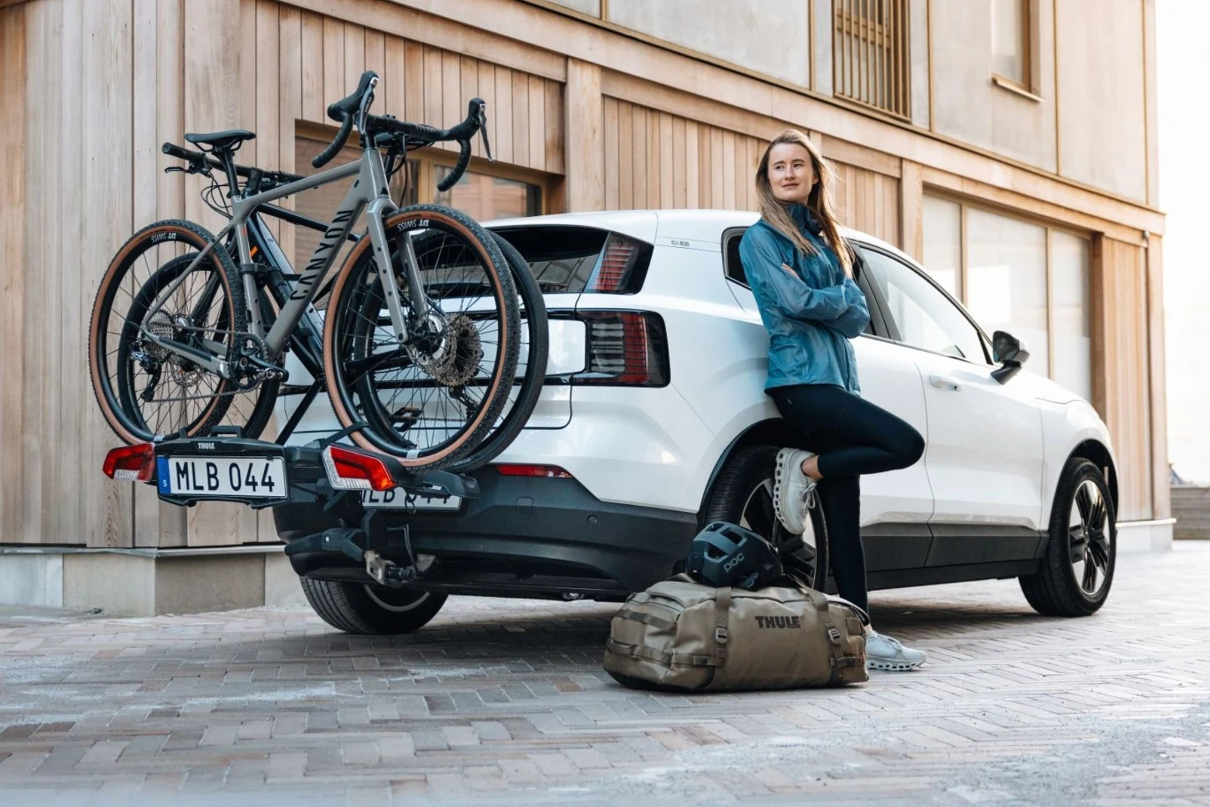 Bagażnik Thule OutPace idealny do ciasnych przestrzeni. Kobieta w sportowym stroju opiera się o biały samochód z zamontowanym bagażnikiem rowerowym, na którym przewożone są dwa rowery. Obok pojazdu stoi sportowa torba i kask rowerowy. Tło stanowi elewacja budynku z dużymi oknami.