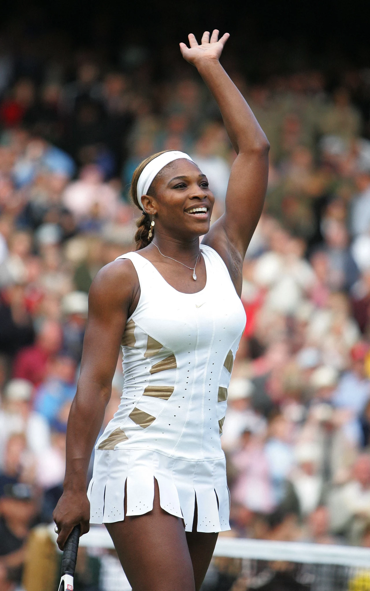 Serena Williams na Wimbledonie, 2004 rok Serena Williams w sportowej białej sukience, uśmiechnięta, unosi prawą rękę w geście powitania lub zwycięstwa, w tle rozmyta widownia na stadionie sportowym.