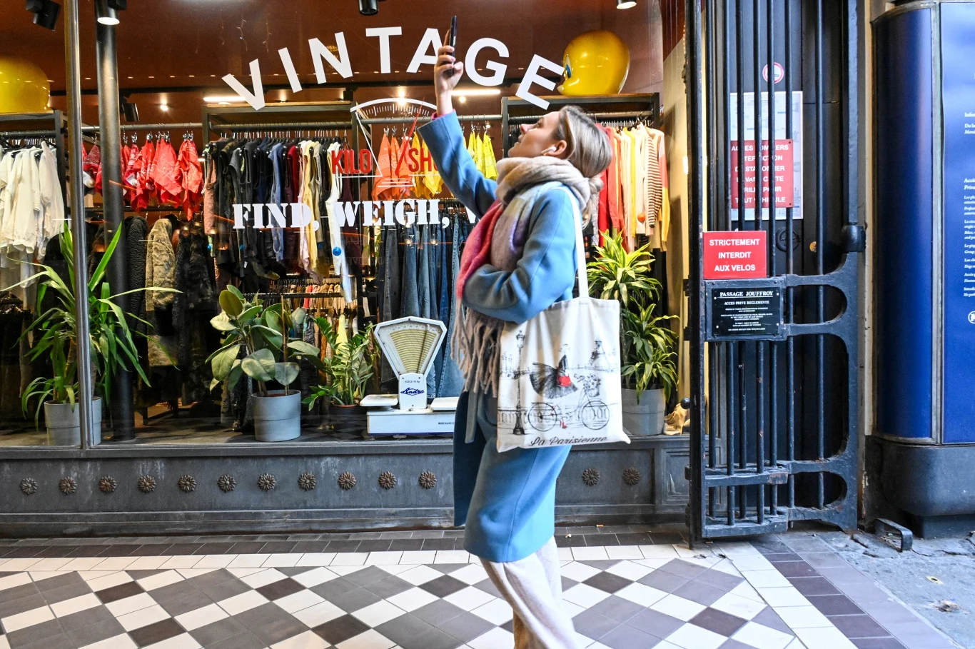 Zetki nierzadko kupują używane ubrania, w związku z czym rośnie popularność "vintage shopów" Kobieta w niebieskim płaszczu ze szalikiem i torbą na ramieniu robi selfie przed sklepem vintage z kolorową odzieżą na wystawie, na tle dekoracyjnych roślin i starych wag.