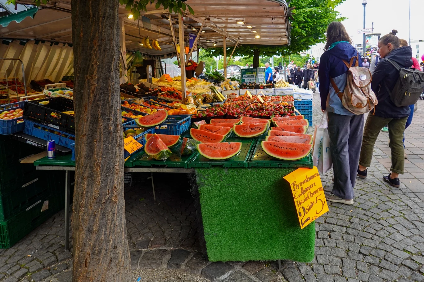 Nawyki zakupowe Zetek znacząco różnią się od nawyków millenialsów, czy boomerów Stoisko targowe z dużym wyborem świeżych owoców, na pierwszym planie arbuzy pokrojone w grube plastry, obok nich inne sezonowe produkty oraz wywieszona żółta tabliczka z ceną, po prawej stronie widoczni przechodnie oglądający asortyment.