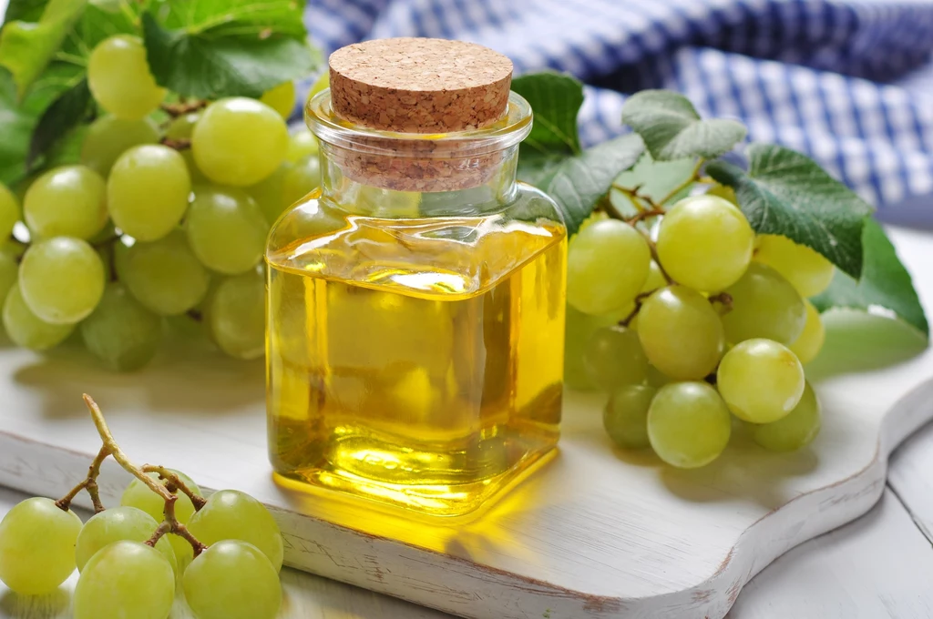 Olej z pestek winogron świetnie pielęgnuje skórę Szklana butelka z żółtym olejem winogronowym zamknięta korkiem, otoczona świeżymi, zielonymi winogronami oraz zielonymi liśćmi, na jasnej drewnianej desce z tkaniną w niebieską kratę w tle.