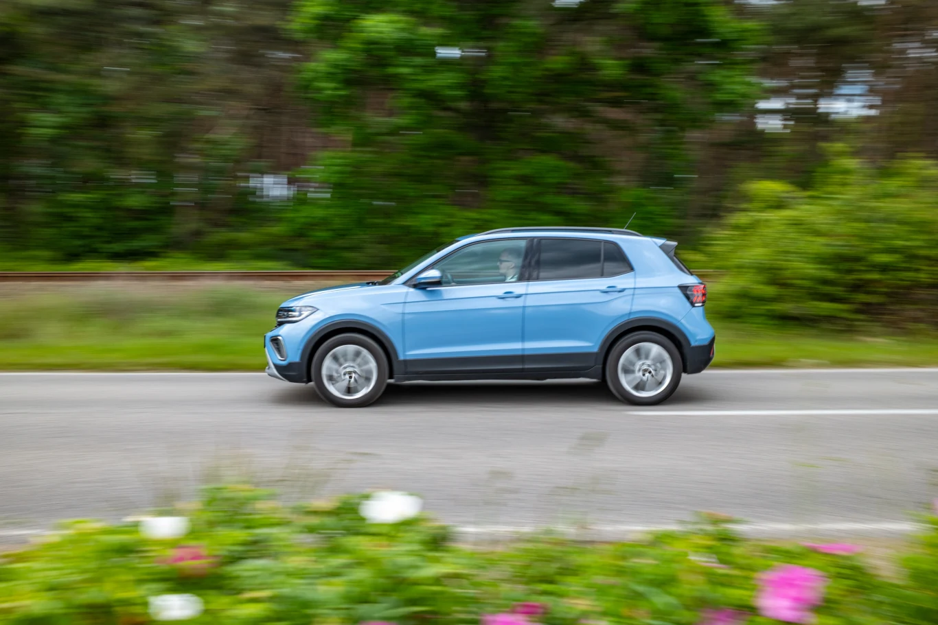 Volkswagen T-Cross ma bardzo “kwadratowe” proporcje; z silnikiem 1.0 TSI o mocy 116 KM jest wystarczająco dynamiczny. Jasnoniebieski samochód osobowy jedzie dynamicznie po asfaltowej drodze, w tle gęsta zieleń drzew i krzewów, na pierwszym planie rozmyte kwiaty.