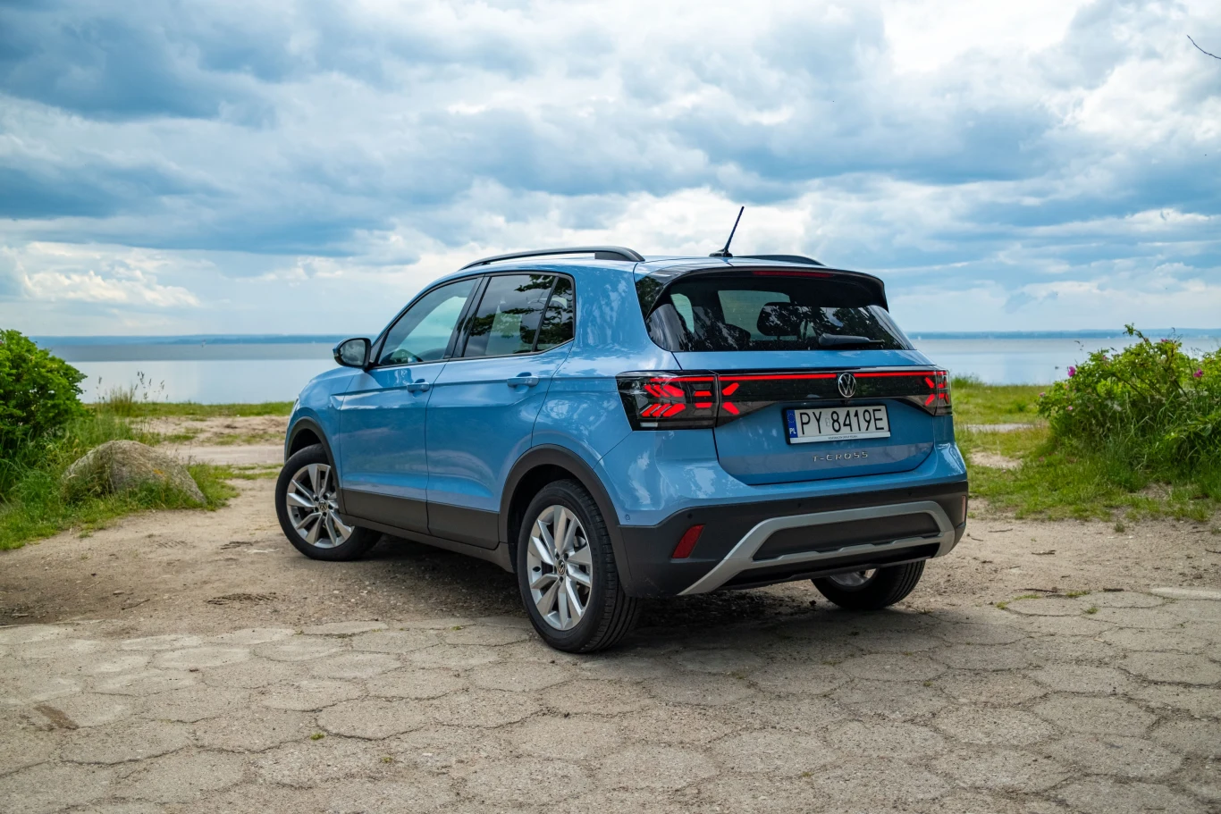 Po face liftingu zmieniły się między innymi tylne lampy. W tej formie T-Cross zostanie z nami jeszcze przez kilka lat. Niebieski samochód SUV marki Volkswagen zaparkowany tyłem na utwardzonym parkingu przy jeziorze lub morzu, w tle widoczne niebo z chmurami oraz zieleń po bokach kadru.