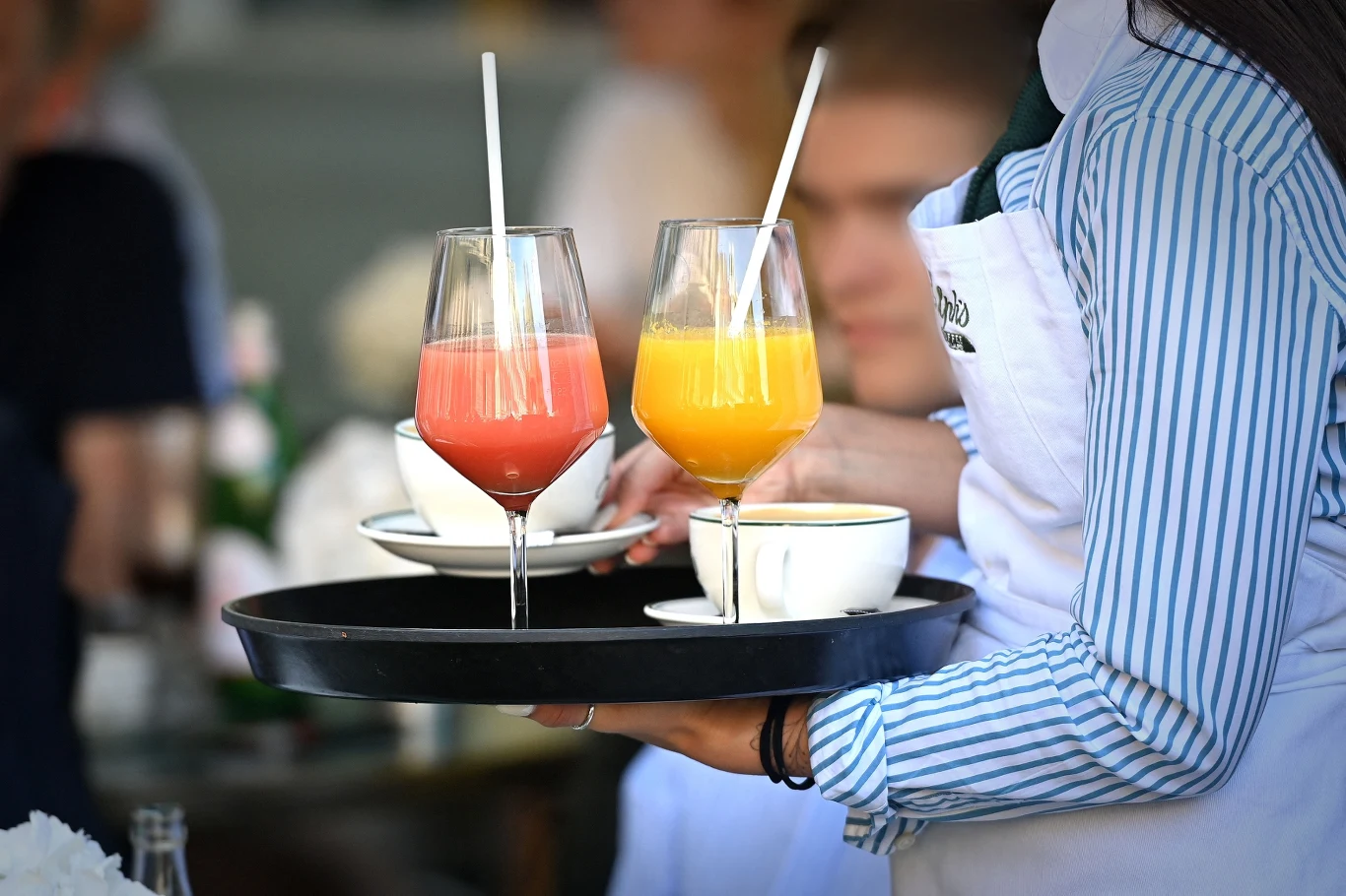 Wśród wydatków, które można ograniczyć młodzi wskazują często jedzenie i picie na mieście Taca trzymana przez kelnera w pasiastym niebiesko-białym rękawie oraz białym fartuchu z dwoma kolorowymi sokami w wysokich szklankach ze słomką oraz dwoma filiżankami z napojami, w tle rozmyci ludzie siedzący przy stole.