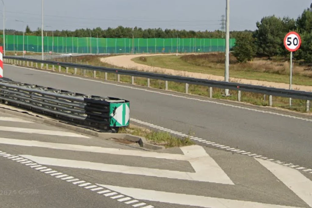 Barierki ochronne przy drodze, znak ograniczenia prędkości do 50 km/h, pas zieleni, ekrany dźwiękochłonne na tle lasu, wyraźne oznakowanie poziome na asfalcie.