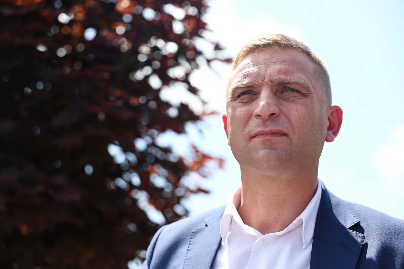 Rzecznik rządu o "aktywistach" Roberta Bąkiewicza Mężczyzna w jasnej koszuli i granatowej marynarce patrzący przed siebie z poważnym wyrazem twarzy, w tle rozmazane liście drzewa i jasne niebo.