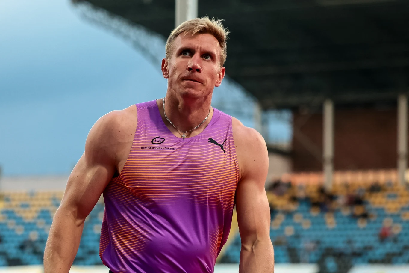 Umięśniony lekkoatleta w fioletowej koszulce sportowej znajduje się na stadionie lekkoatletycznym; w tle widoczne są trybuny z niewyraźną publicznością.