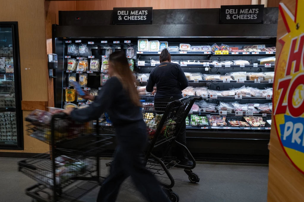 Zamiast gotować niektórzy młodzi lubią kupować gotowe dania lub diety pudełkowe Dwie osoby robiące zakupy przy lodówkach z wędlinami i serami w supermarkecie, jedna z nich przegląda produkty, druga przejeżdża z wózkiem obok regałów.