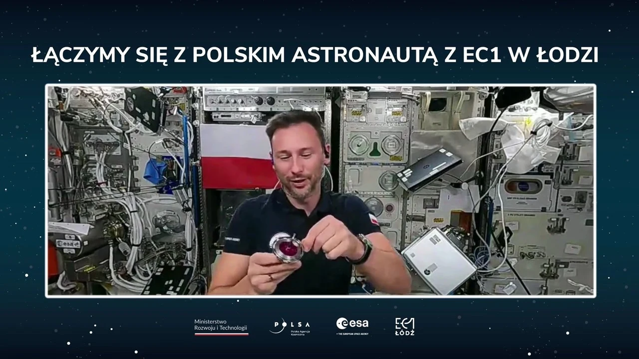 Astronauta w stroju roboczym unosi się w module stacji kosmicznej, trzymając mały przedmiot w dłoniach, a za nim widoczna jest polska flaga oraz panele i urządzenia laboratoryjne.