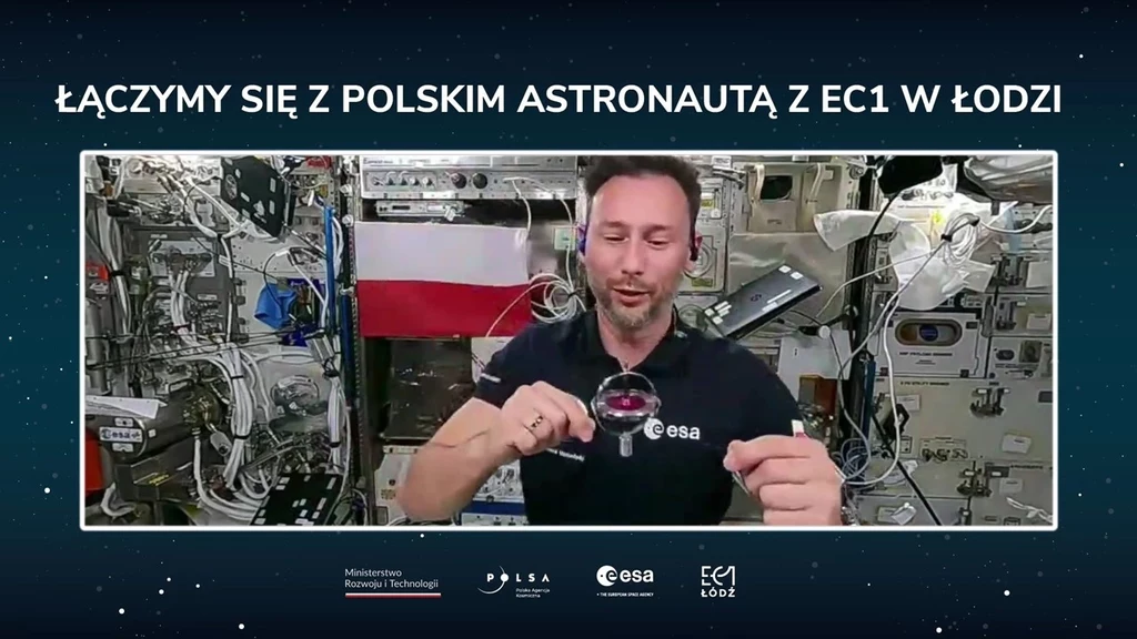 Polski astronauta unoszący się w przestrzeni Międzynarodowej Stacji Kosmicznej, w tle polska flaga oraz aparatura badawcza i urządzenia stacji kosmicznej.