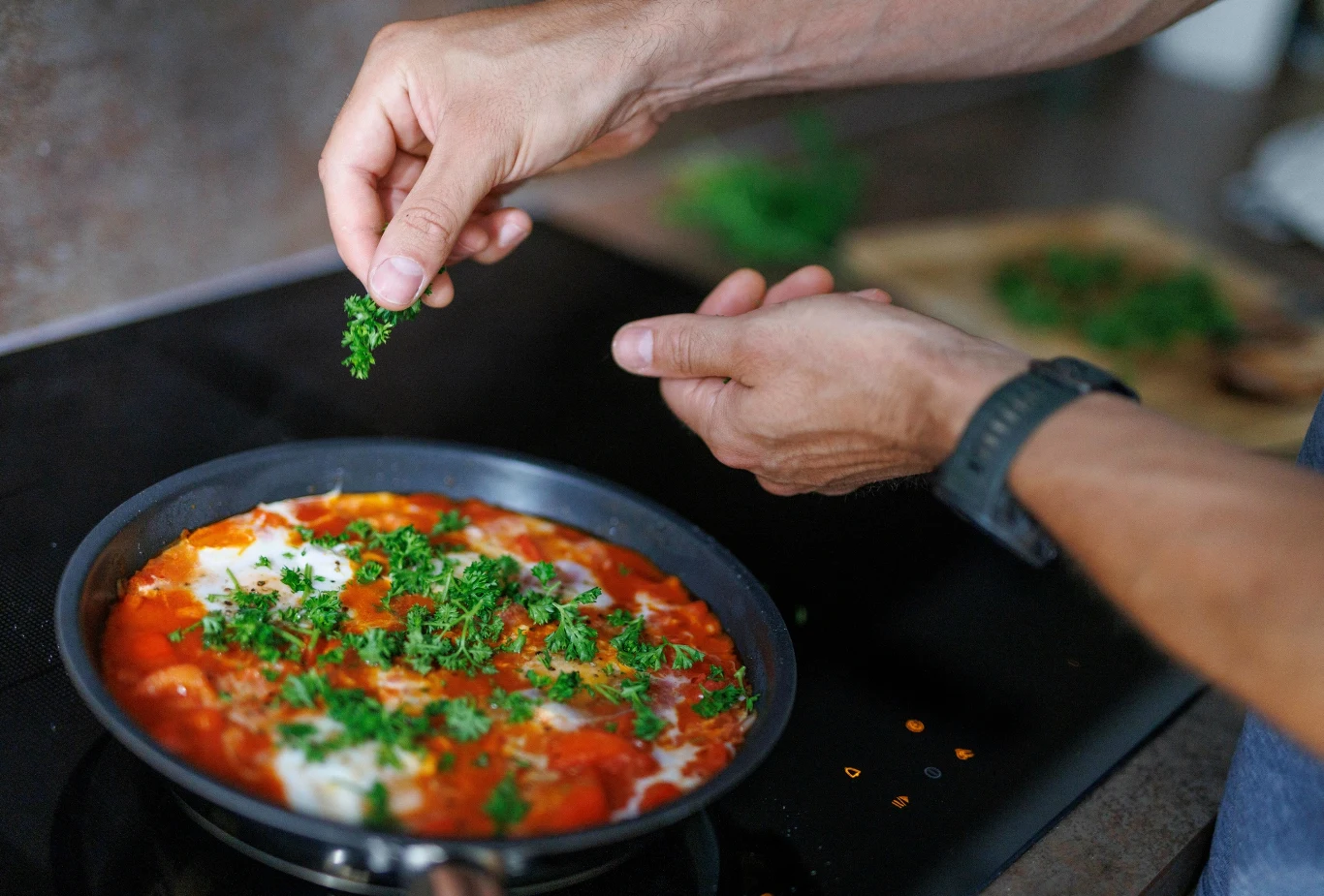 Przygotowywanie posiłków w domu to dla Zetek dobry sposób na dodatkowe oszczędności Dłonie osoby dodającej świeże, posiekane zioła do patelni z pomidorowym sosem, na którym widać również jajka gotujące się na wolnym ogniu.