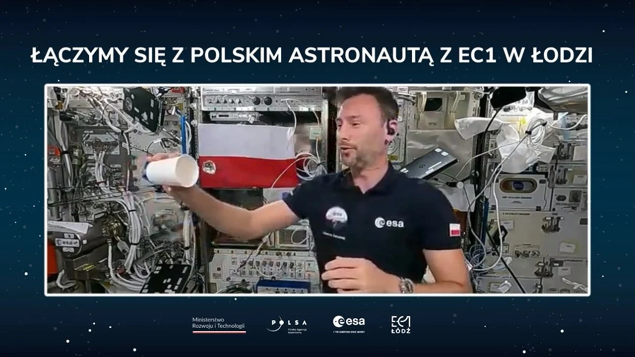 Polski astronauta znajduje się w stacji kosmicznej otoczony licznymi urządzeniami elektronicznymi i przewodami, trzyma w ręku biały kubek, w tle widoczna jest flaga Polski oraz oznaczenia ESA i partnerów projektu.