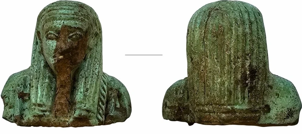 Metalowa statuetka przedstawiająca egipską głowę z długimi włosami, pokrytą patyną; widok z przodu uwydatnia uszkodzenia twarzy, natomiast z tyłu widoczna struktura fryzury.