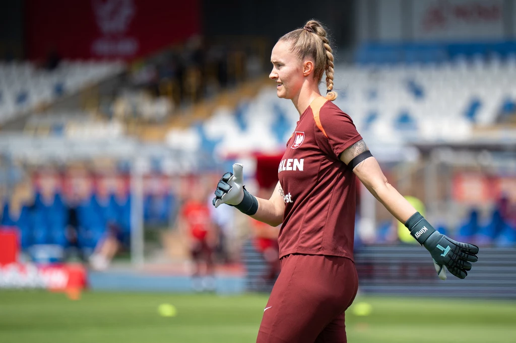 Kobieta w stroju bramkarki piłkarskiej biegnie po murawie stadionu, na rękach ma rękawice bramkarskie, w tle rozmazane trybuny z niebieskimi i białymi siedzeniami oraz inni ludzie.