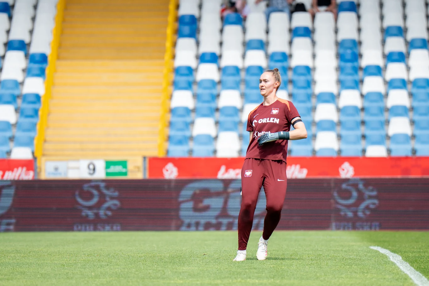 Kinga Szemik Piłkarka ubrana w bordowy strój sportowy biegnie po zielonej murawie stadionu, w tle puste rzędy niebiesko-białych krzeseł oraz fragment żółtych schodów.