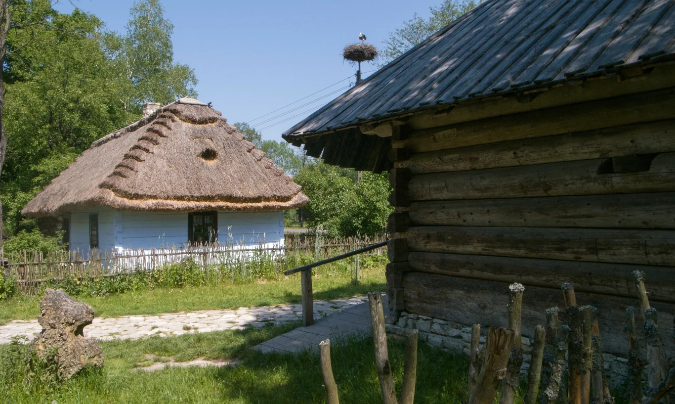 Drewniana chata z dachem krytym strzechą obok białej, zabytkowej chaty w otoczeniu zieleni, z drewnianym płotem i bocianim gniazdem na słupie.