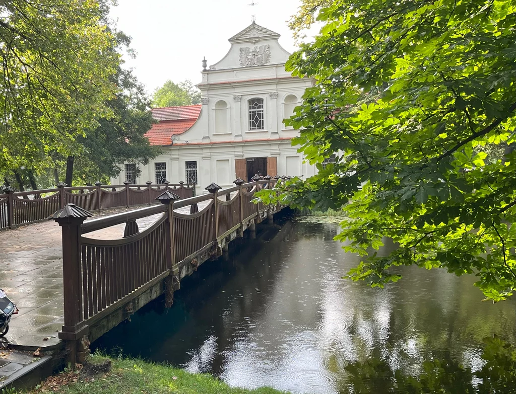 Most drewniany prowadzący nad wodą do historycznego budynku o białej elewacji z barokowymi zdobieniami, pośród otaczającej zieleni drzew. Odbicie fasady w lekko wzburzonej powierzchni stawu.