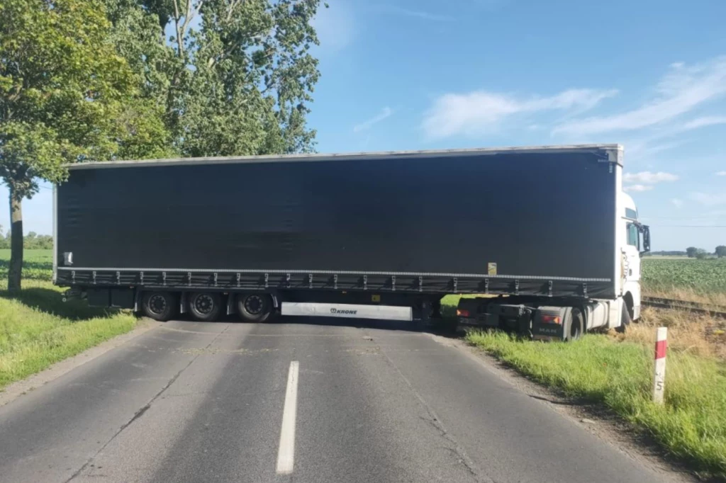 Ciągnik siodłowy z naczepą zablokował całą szerokość drogi, uniemożliwiając przejazd innym pojazdom, wokół widoczne pola i drzewa, a asfaltowa jezdnia przebiega przez wiejski krajobraz.