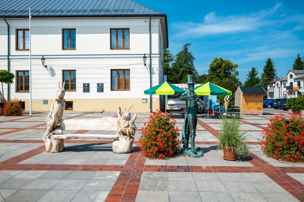 Rzeźby przedstawiające postacie bajkowe ustawione na placu z brukowaną nawierzchnią, na tle białego budynku z kilkoma oknami, wokół kolorowe kwiaty w donicach i zielone stoiska handlowe.